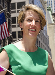Woman with short brown hair and green sleeveless dress smiling into camera with United States flag in left background