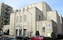 The view of a low-rising, setback gray building with a multi-story entrance decorated with geometric shapes.