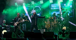 A band all dressed in dark clothing performing on stage; a singer with a white LED head-dress, two guitarists, a keyboardist, and a bongo player are seen behind fog coloured green from the stage lighting