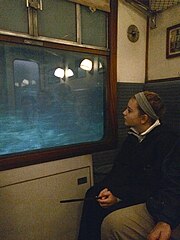 Inside a 1950s-style train compartment. On the left is a window with a projected blue-green image behind it. On the right is a person watching the projection through the window. The compartment lights and sliding door are partially reflected in the glass.