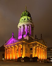 Nachtfotografie von einem klassizistischen Gebäude mit Säulen und einem hohen Kuppelturm mit goldener Figur an der Spitze. Von oben nach unten ist das Gebäude in den Farben Grün, Violett und Gelb beleuchtet.