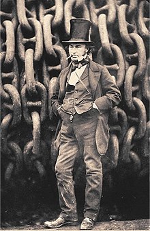19. yüzyılda dev demir zincirlerin önünde duran, üzerinde ceket, pantolon ve yelek olan, silindir bir şapka takan, ağzında sigara, elleri ceplerinde bir adam.