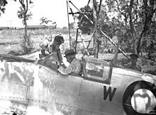 An old man wearing a hat and white shirt sitting in the cockpit of a silver aircraft. A younger man is leaning into the cockpit.