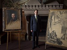 George Friedrich Prinz von Preussen poses standing and in a suit between two paintings.