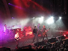 Powderfinger are on stage, with Coghill at left on drums, then Middleton on guitar, Fanning mid-stage singing into a microphone, Collins on his bass guitar and Haug at extreme right.