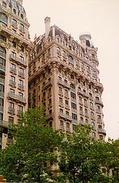 The Ansonia as seen from across Broadway. The facade is made of limestone, and there are many windows with iron balconies. In the foreground are trees.