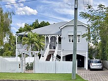 A long shot of a high-set Queenslander residential house made of timber with a corrugated iron roof. An external staircase leads to a veranda on the upper level of the house, and a white picket fence sits at the front of the property.