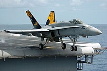 F/A-18 Hornet fighter departing aircraft carrier. A gray aircraft, with blue and yellow fins, has just left the edge of carrier's deck, as evident through the extended landing gear.