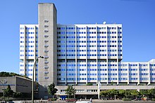 Frontale Farbfotografie des modernen, vierzehn-stöckigen Bürogebäudes, das aus weißer Fassade und vielen kleinen Fenstern besteht. Im linken Bereich ist ein grauer Turm mit kleinen länglichen Fenstern.