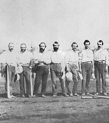 Seven members of the New York Knickerbockers baseball team standing on a field. They are wearing white shirts and dark pants.