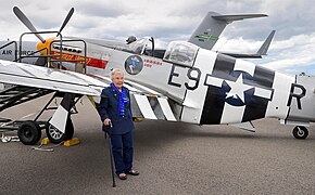 Olsen, using a cane for support, standing in front of a P-51 Mustang on static display. The airplane is painted in [[Invasion stripes]].