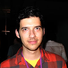 Vreeland's face is close-cropped and bright with a harsh flash. He is in a very dark, indoors setting, and wears a red and black plaid shirt. He is white and has short, dark hair, some stubble, and no glasses.
