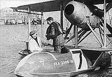Photograph of a man in a flight suit and life preserver, standing the cockpit of a biplane floating on water. He is speaking to another man stood just outside the aircraft.