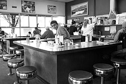 Black-and-white photograph of a restaurant's interior, with three people behind a counter serving patrons seated at a curved counter