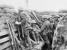 Several members of the 1/7KR in a deep trench. In the foreground, another member stands on a firing step peering over the parapet composed of sandbags.
