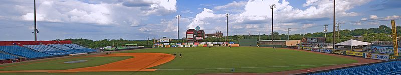 A panorama of the ballpark taken from the right field seats showing the field, entire outfield wall, guitar scoreboard, green trees outside the park, and a partly cloudy blue sky above