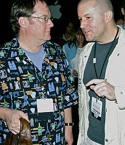 Jonathan Ive is pictured to the right, talking to John Lasseter who is pictured to the left. The Apple logo can be seen in the background.