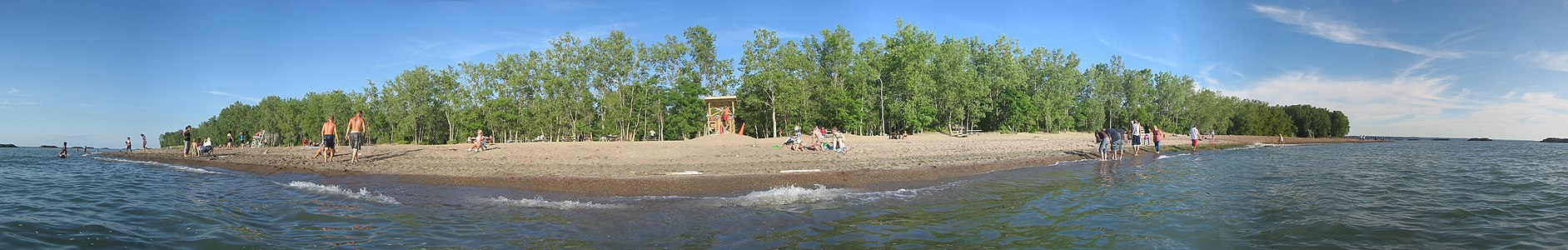 A view of a sunlit beach, that is flanked by green trees, from the water under a mostly clear, blue sky with several vacationers.