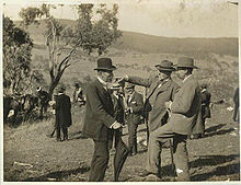 Tiga lelaki pertengahan umur dengan janggut pendek dalam sut formal dan topi berdiri di padang berbukit terbuka dengan pokok tunggal berdekatan