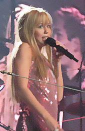 A mid shot of a female performer, wearing a long blonde wig, singing into a microphone. She is wearing a dress with pink sequins. A screen projecting images of the performance appears behind her.