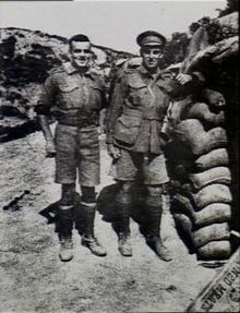 Two men in military uniform standing side-by-side. The man on the left is slightly shorter and stockier, and the man on the right has his arm leaning on a stacked up pile of sandbags.