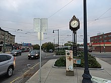 This image is an October 2021 view of the Boston Neighborhood, Cleveland Circle from the intersection of Beacon Street and Chestnut Hill Avenue. These cross The image is looking northeast down Beacon Street. Prominent landmarks shown in the photograph are the Cleveland Circle neighborhood clock and the Cleveland Circle Station, which serves as terminus of the MBTA C line. It also shows the primary commercial drag of the neighborhood, which goes though this portion of Cleveland Circle.