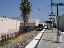 A train station at ground level with a track on the left side. A light rail train is stopped at the station. Various buildings are located to the left of the station.