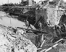 The remaining column of a destroyed bridge, with surrounding rubble that has partially blocked the flow of a canal. Members of the 2/5LF walk across some of the rubble.