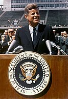Presiden Kennedy is pictured speaking behind a podium. Rice University's stadium is visible behind him.