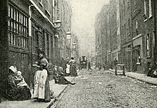 A street in Edwardian London with shop fronts and tall terraced buildings; women and children are in the foreground