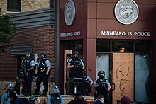 A half-dozen officers wearing light blue shirts, black gas masks and black bullet-proof vests, carrying long tear gas launchers, standing in front of a corner brick and glass building with boarded up windows, identified with the seal of Minneapolis and "Minneapolis Police" in large white letters