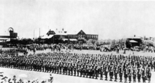 Groupe de soldats avec leur fusil à l'épaule sur un terrain de parade entouré d'une large foule