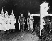 A black-and-white image picturing a group of white people robed in Ku Klux Klan regalia in a field next to a burning cross