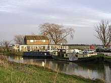 River scene of two-storey wooden building with chimney with Fish and Duck written on the front wall some canal long-boats in the foreground