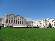 A drive leads past a manicured lawn to large white rectangular building with columns on it facade. Two wings of the building are set back from the middle section.
