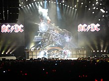 Quatre membre de groupe sur scène devant un public, avec une locomotive factice et deux logos AC/DC en fond.