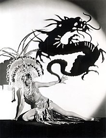 Photographie en noir et blanc montrant une femme agenouillée sur scène devant l'ombre portée d'un dragon.