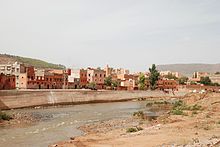 A river runs across the image from left to right with a town in the background, behind a concrete flood defence. The foreground shows a stony, sparsely vegetated river bank.