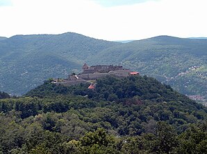 A castle atop a forested hill, surrounded by mountains.