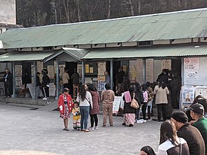 A line of about 15 women leads out from the entrance to a building with a sheet metal roof and information posters covering the walls. A shorter line of men leads out from another door, while other people wait around the sides.