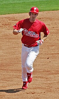 A man in white pants, a red baseball jersey with "Phillies" on the chest, and a red batting helmet with "P" on it runs on a dirt surface.