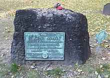 A rectangular, rough-hewn block of stone on the ground. There is a walkway in the foreground, grass and trees in the background. A weathered plaque on the stone reads: "Samuel Adams, Signer of the Declaration of Independence, Governor of this Commonwealth, A Leader of Men and an ardent Patriot."