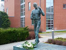 A bronze statue of Jobs with a green patina. Jobs is holding a remote control and gesturing as though in the middle of a presentation. Flowers are placed at his feet.