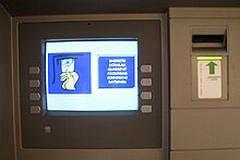 Frontale Farbfotografie von einem Geldautomat. Auf dem Bildschirm ist links eine Grafik mit einer Hand, die die Karte in den Schlitz schiebt. Rechts davon steht in Latein „Inserito scidulam quaeso ut faciundam cognoscas rationem“.