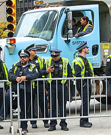 A group of Chicago police officers behind a fence at a roadblock. A Chicago public works truck, also used to block the road, is behind the officers. One of the officers is looking at his cell phone.