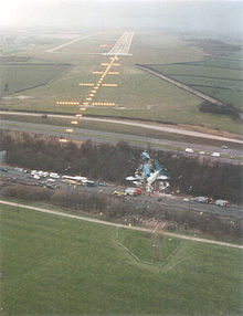 The wreckage of an aircraft lies on ground between two road carriageways running left to right. The runway with landing lights lit is several hundred metres further away.