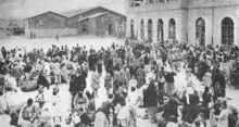 Large group of people gathered in a town square, holding some possessions