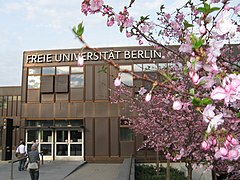 Frontale Farbfotografie eines dunkelbraunen Gebäudes mit der Aufschrift „Freie Universität Berlin“. Das zweigeschossige Gebäude wird rechts teilweise von einem Kirschblütenzweig verdeckt.
