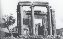 A frontal black and white picture of ionic temple ruins showing a massive entrance door and a smaller side door. Two children in head covers sit at the right of the temple facade.