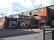 A red-bricked building with a blue sign reading "GREENFORD STATION" in white letters and people in front all under a blue sky with white clouds